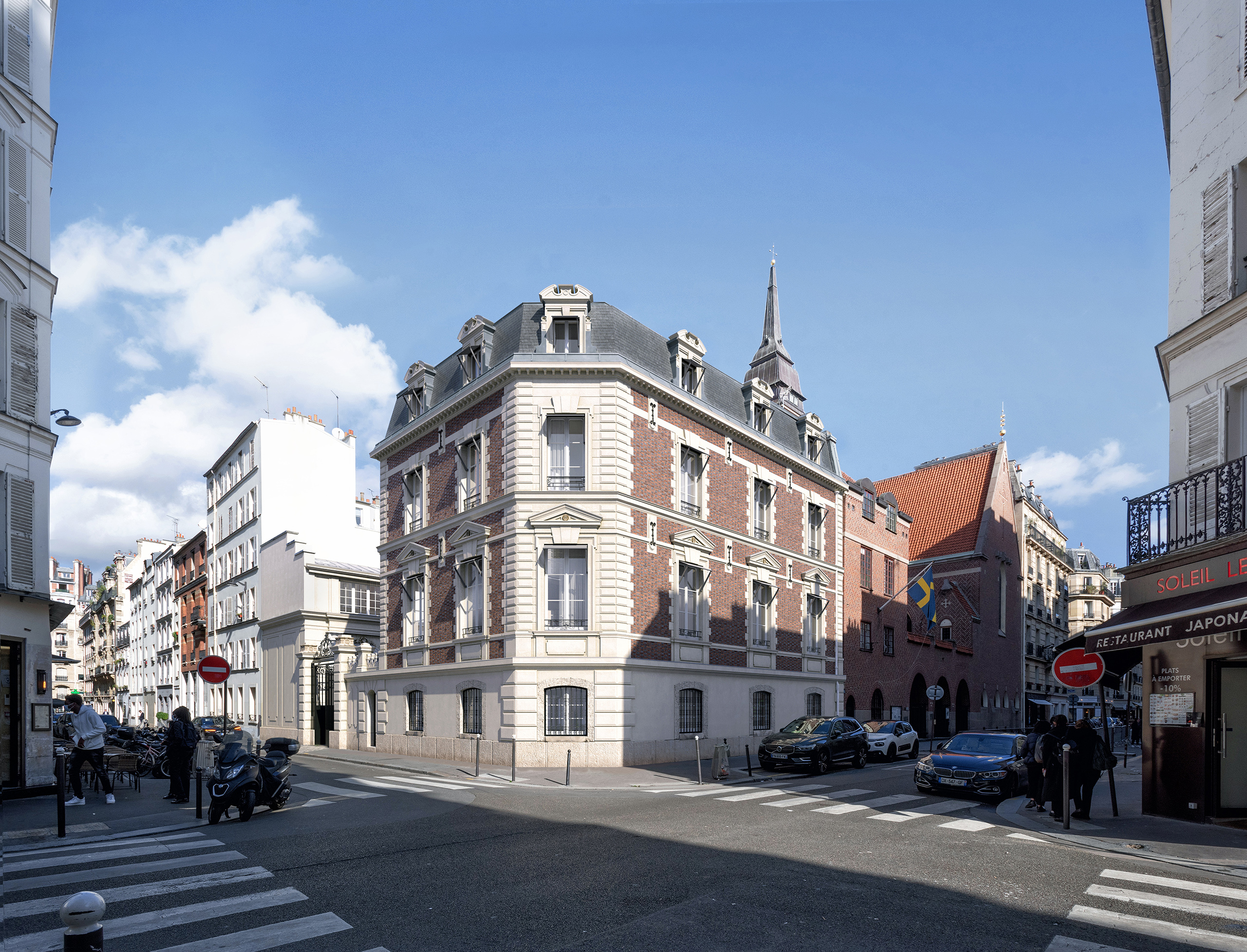 Jost – immeuble en briques rouges et pierre de taille à l'angle d'une rue parisienne, toiture mansardée, ciel bleu
