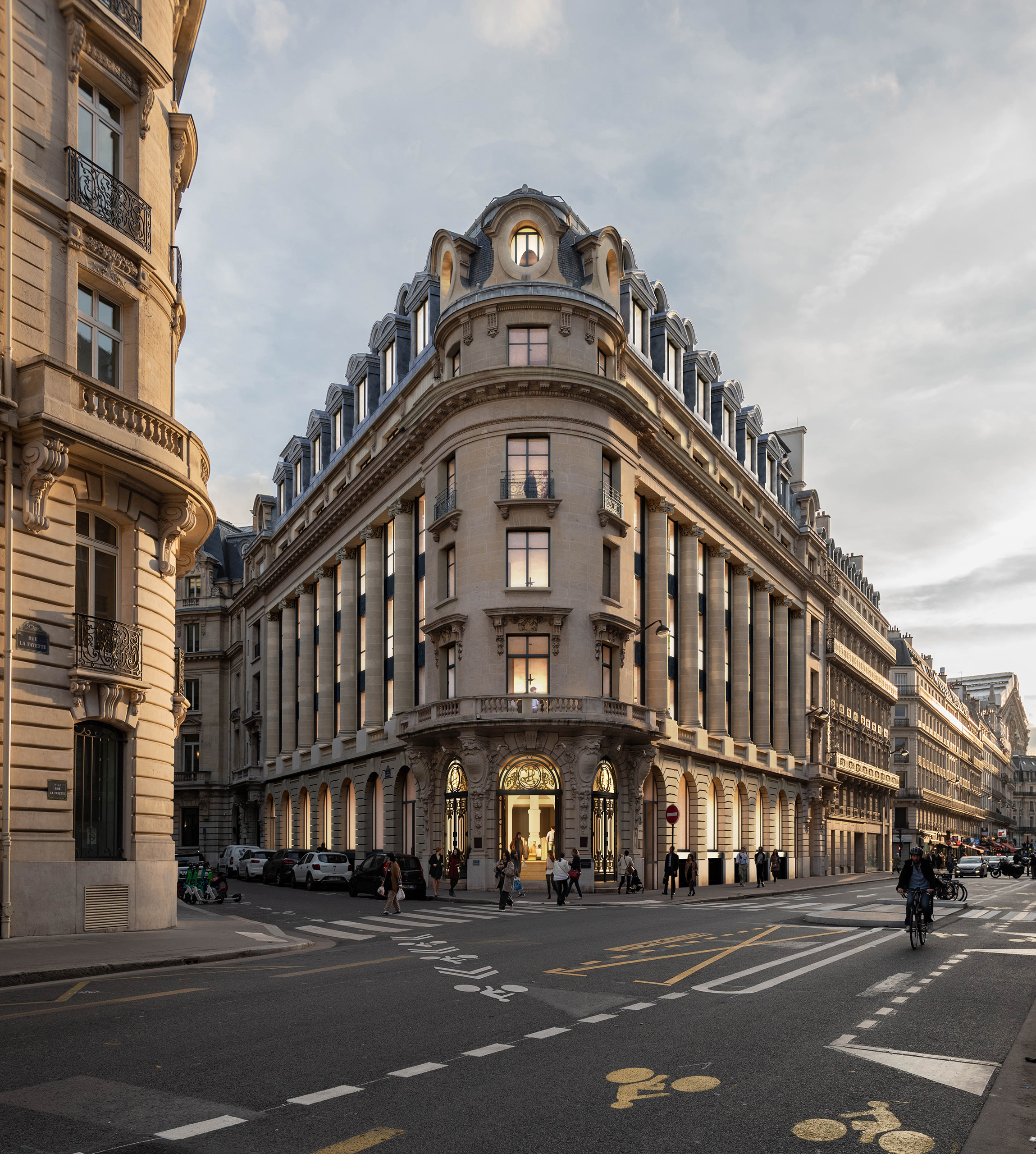 Angle du projet La Fayette au crépuscule – immeuble haussmannien en pointe avec dôme mansardé, entrée dorée et insertions vitrées noires, avenue de l'Opéra Paris
