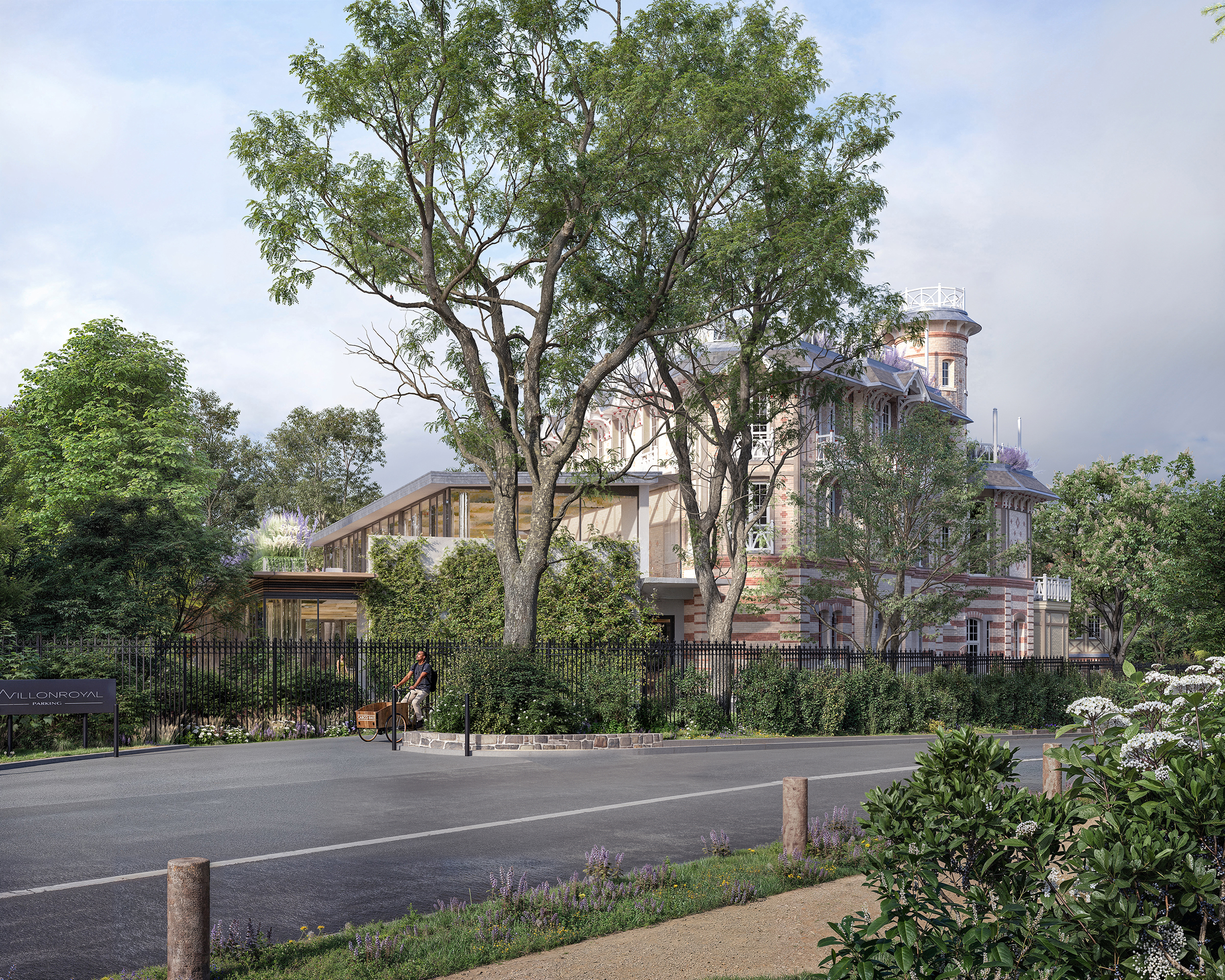 Vue depuis la rue sur un ensemble de bâtiments historiques en briques bicolores et tour d'angle, extension contemporaine vitrée et végétalisée, clôture en fer forgé, cycliste au premier plan, végétation dense